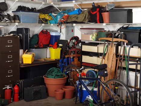 An indoor storage space or garage filled with various household and gardening items, including plastic storage bins, some in clear, blue, and black, stacked on wire shelving units along the back wall. Several toolboxes and containers in different colours, such as a yellow and some red bins, are visible on a work surface. A green garden hose is coiled in a large terracotta pot in the foreground, accompanied by empty smaller pots. A blue and orange bicycle leans against a wooden support structure, with some tools and equipment nearby. The wall-mounted shelving holds additional items, including a black toolbox and a red bag, with cables and extension cords hanging loosely. The overall scene suggests a cluttered but organized space used for storage and disposal preparation, typical of a property in Tower Hamlets, with a variety of textured plastic, metal, and wooden objects. The lighting appears to be from an overhead source, illuminating the different objects and surfaces clearly, echoing common storage areas requiring rubbish removal services from Commercial Waste Tower Hamlets.
